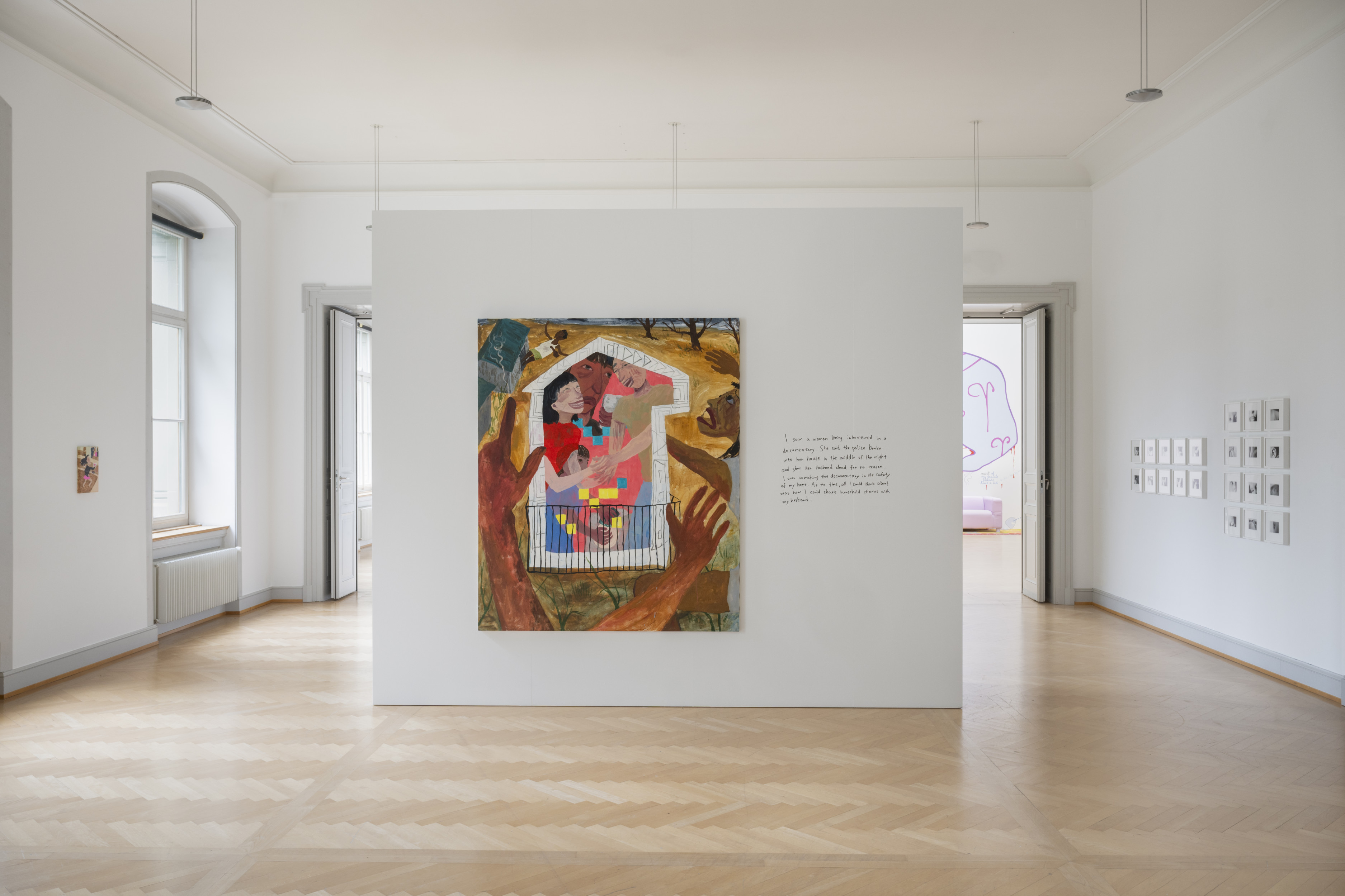 A contemporary art gallery with minimalistic, white walls and light wooden floors. In the center, a large, colorful painting is displayed on a freestanding white wall. The artwork features four intertwined human figures at the center of the image, against a bright pink background. The figures are enclosed by a thick white line resembling a house. Surrounding the composition at the center of the frame is a barren landscape of ocre and taupe.Arts reach toward the center of the image from the lower foreground of the image. On either side of the image, distressed looking figures wave their hands. To the right of the painting, handwritten text is visible on the wall.