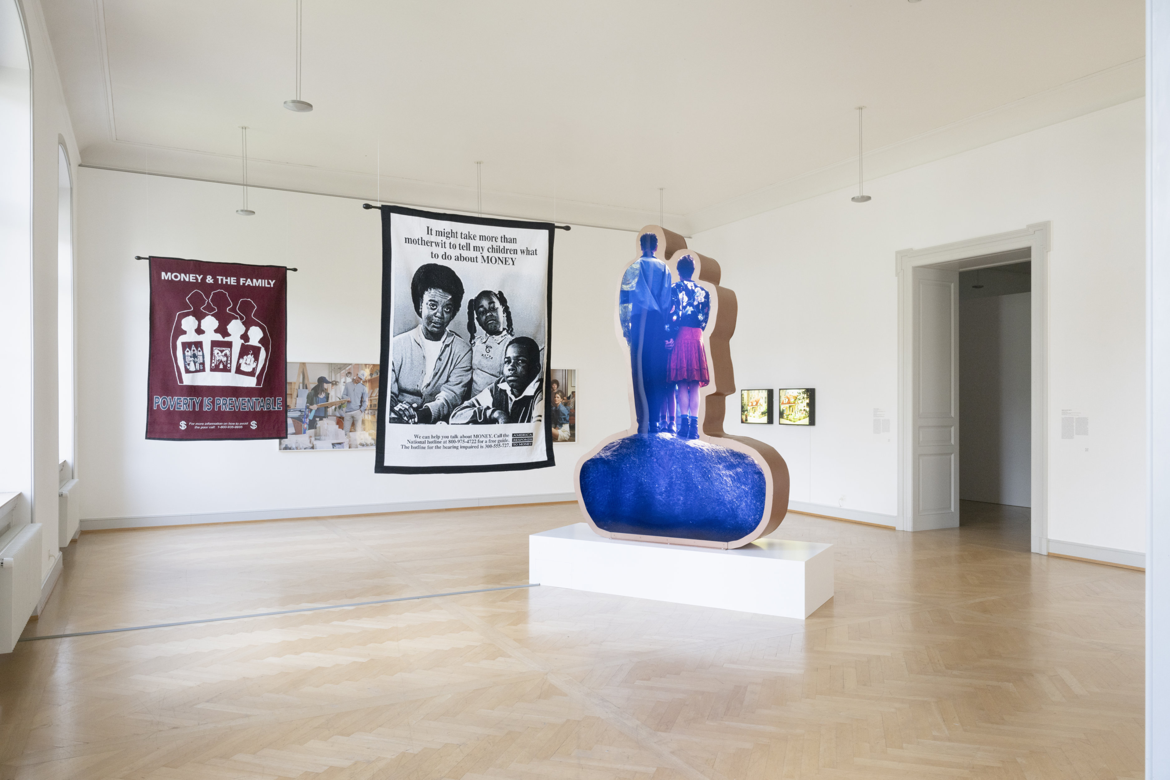 A contemporary art gallery featuring hanging textile works and a freestanding sculpture. On the left, a maroon textile reads "MONEY & THE FAMILY: POVERTY IS PREVENTABLE," and a black-and-white textile displays a Black family portrait with text. In the center, a three-dimensional photographic cutout, showing the back of two figures, and the obscured legs of a child.
