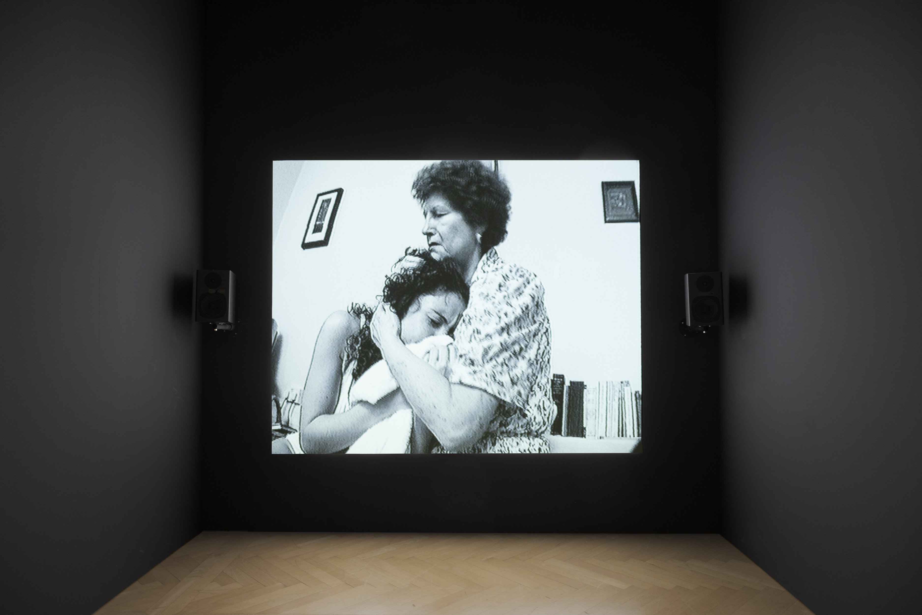 A darkened gallery with a video projection on the center wall, showing an older woman cradling the head of a younger adult. Both have their eyes closed. The image onscreen is grainy.