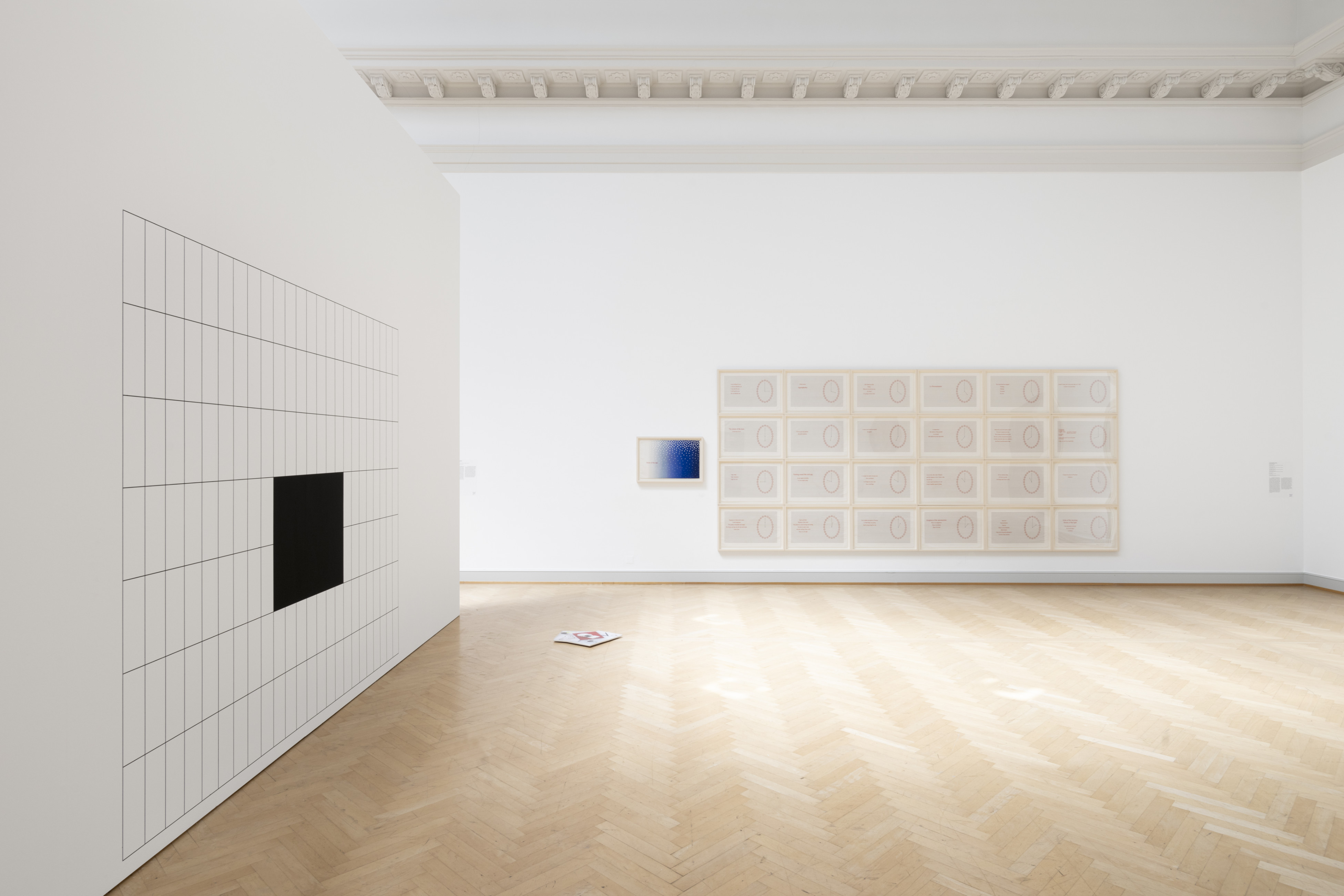A contemporary art gallery, with light wooden floors displays two large wall works. On the left, a grid divided up into large rectangles, with a filled in section in black at the middle of the grid. On the far wall, separately framed images, closely hung, also form a grid. Each images features a circle in red, with text on the left. TO the left of this composition is a small framed image made up of a blue gradient. .
