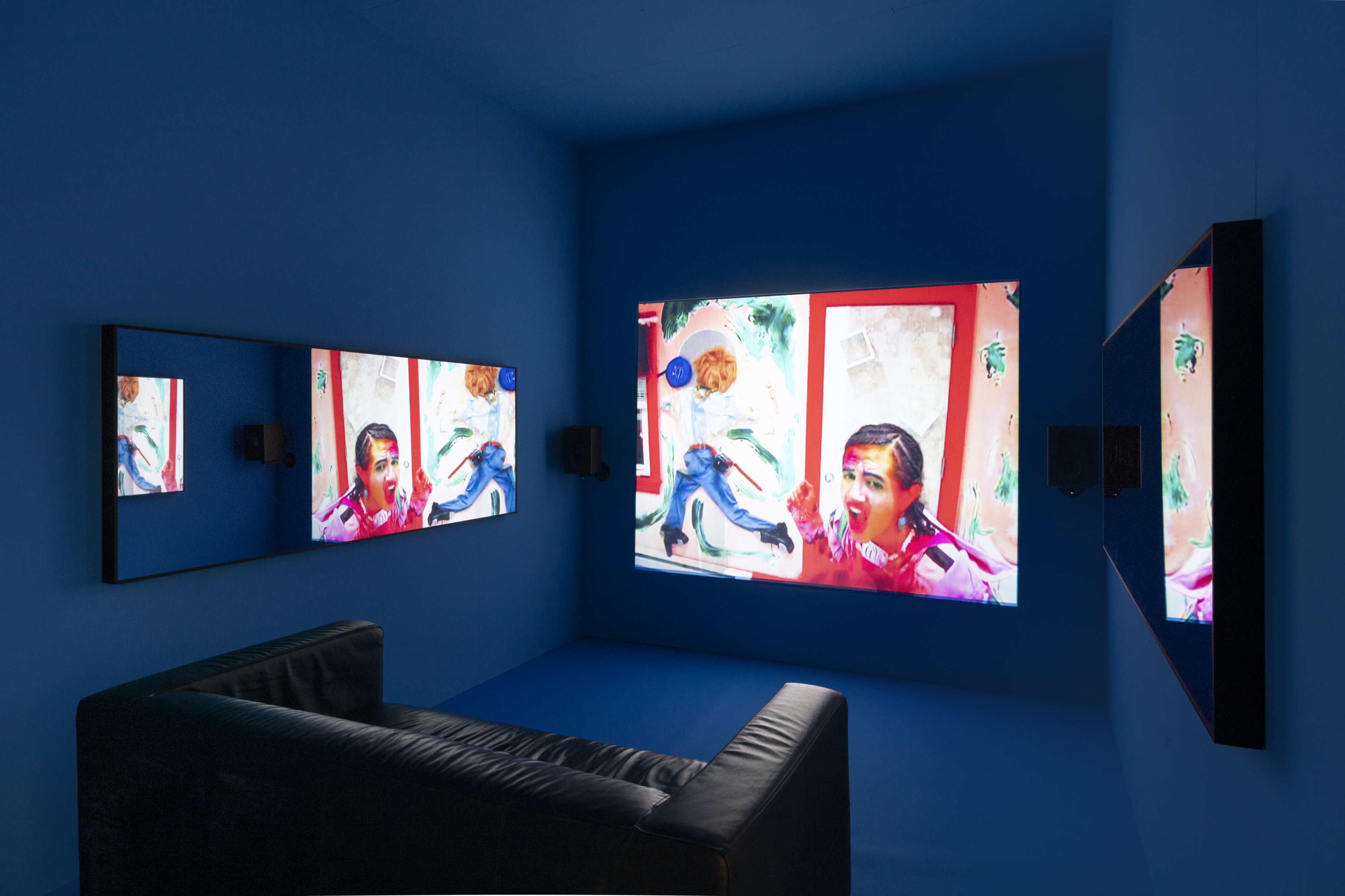 A dark blue room with leather couch facing a large video projection on the wall, displaying a colorful scene featuring a person in facepaint and exaggerated experssions. Two mirrors on the side walls reflect the video.