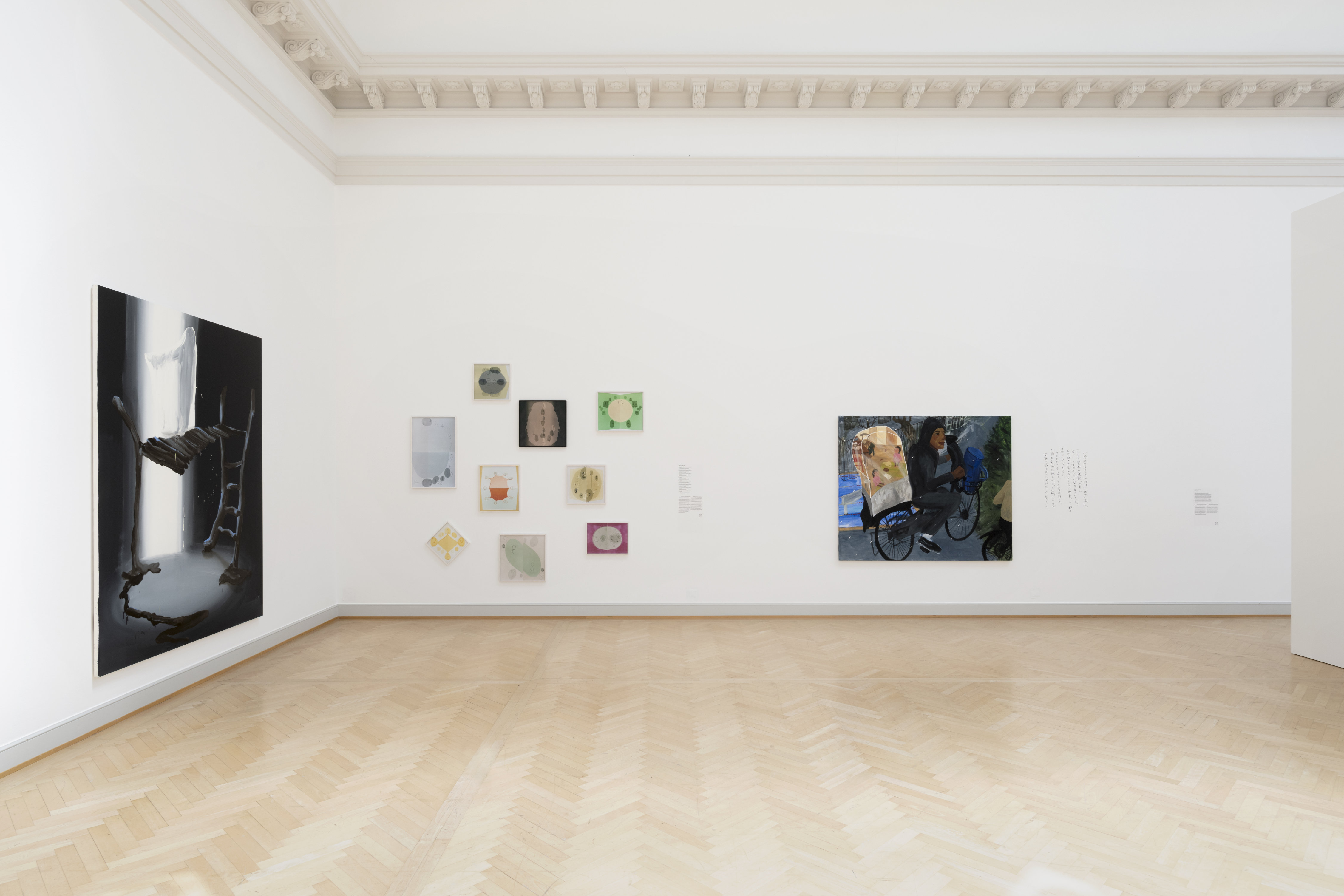 The corner of a bright contemporary gallery with light wood floors. On the left wall a large photo-realistic painting. On the adjacent wall, 9. small, colorful canvases with circular, clock-like forms at the center. On the right is another large canvase, featuring a figure depicted in 3/4 profile, riding a bicycle and carrying a large, light-colored package on the back.