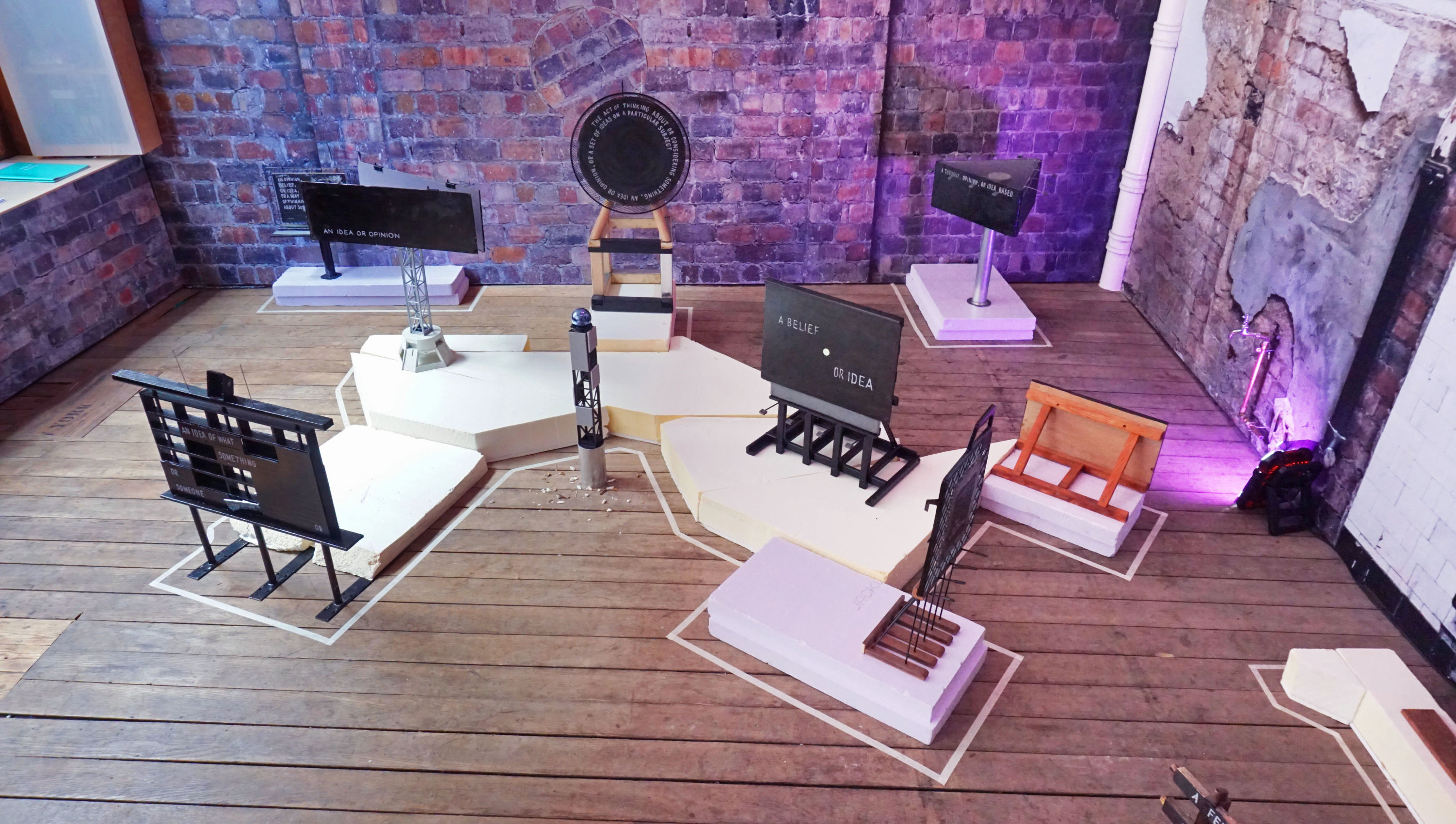 An overhead shot of a brick-sided gallery with wood-plank flooring. Objets are assembled atop white foam platforms and surrounded by a white line of masking tape. Purple light shines on the floor and far wall.