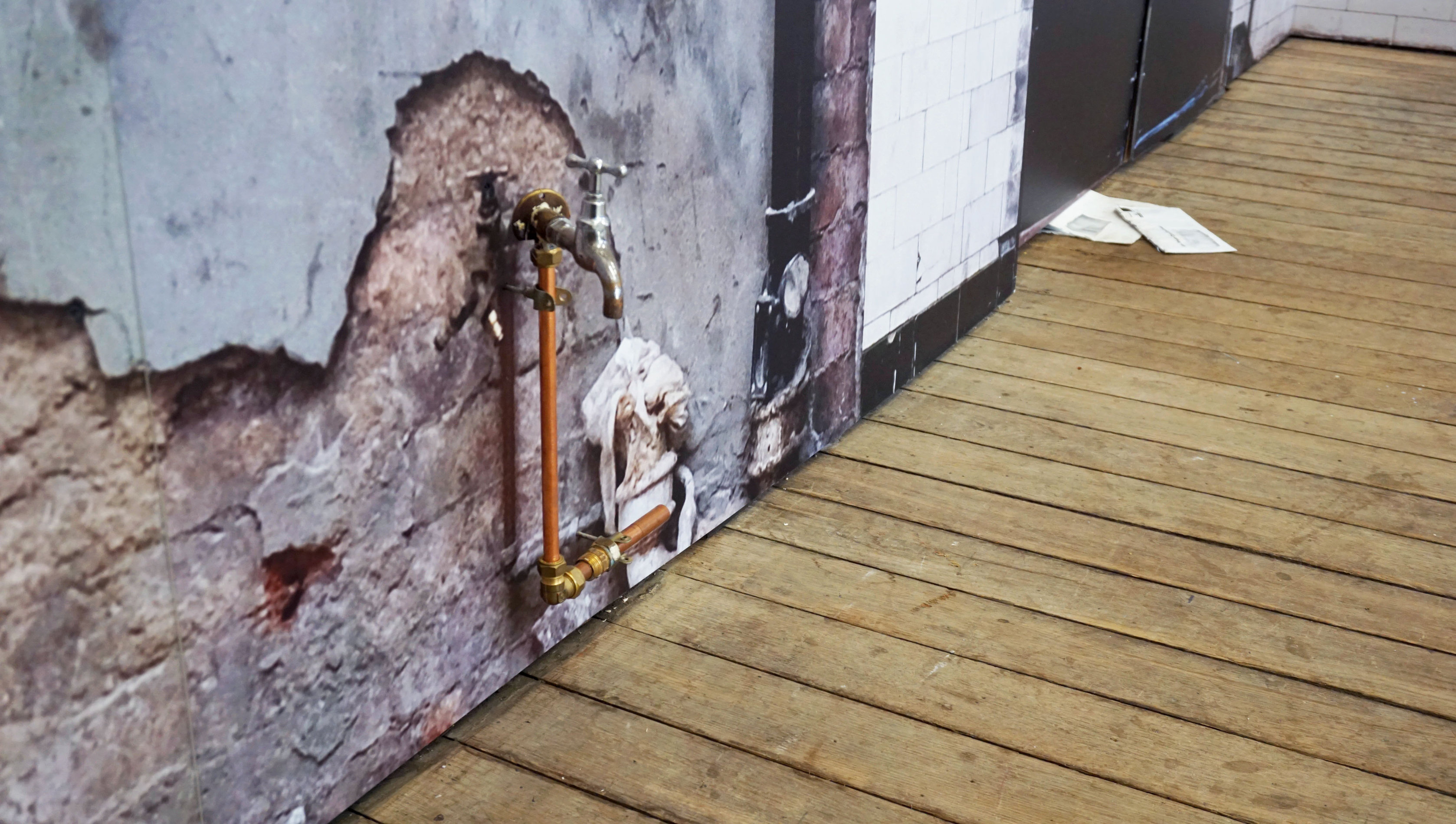 A closely-cropped image of a wall and wood plank floor. The wall consists of broken concrete atop which a water tap is fastened. The texture of the wall is flat, an appears partially photographic, with the water tap appearing three-dimensional.