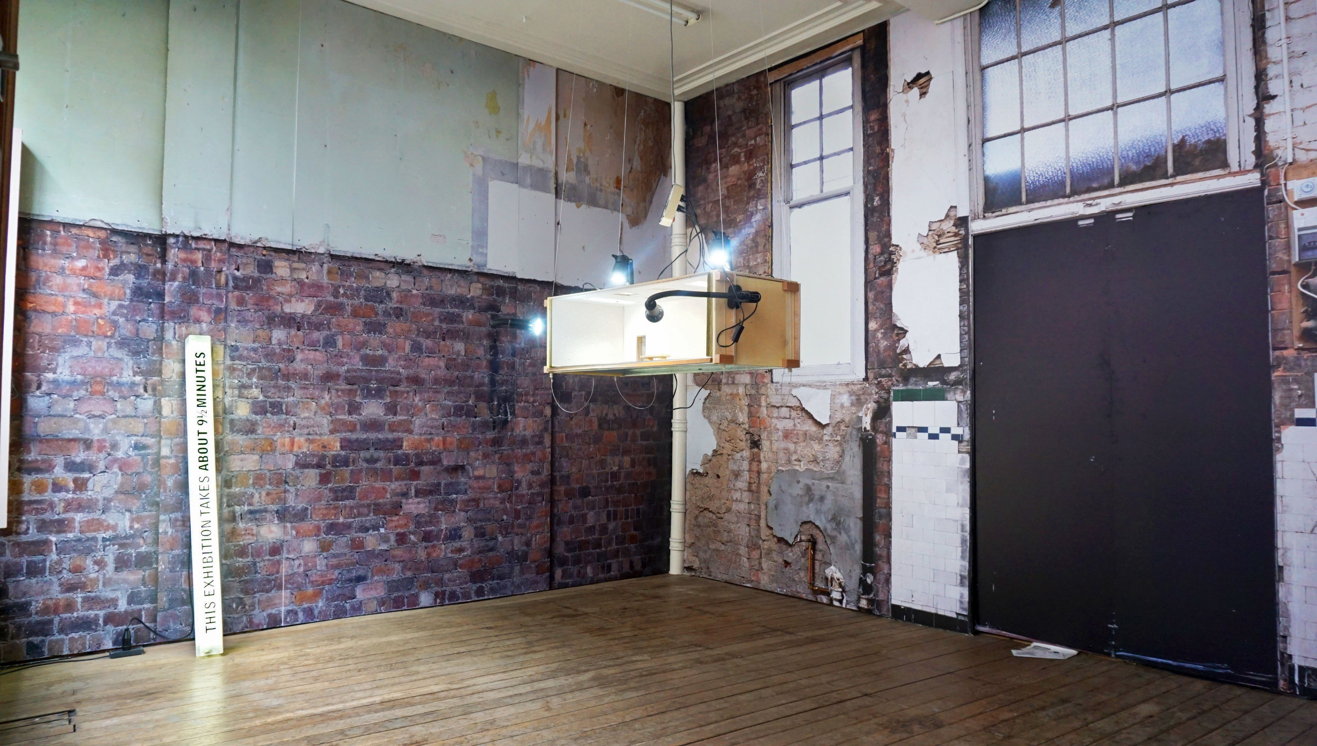 A small brick-lined gallery with high ceiling and tall frosted glass windows. From the ceiling is suspended a large, low-hanging wooden box with liughts attached. These lights shine on a wooden plant leaning vertically against the far wall with the words "THIS EXHIBITION TAKES ABOUT 9 1/2 MINUTES"