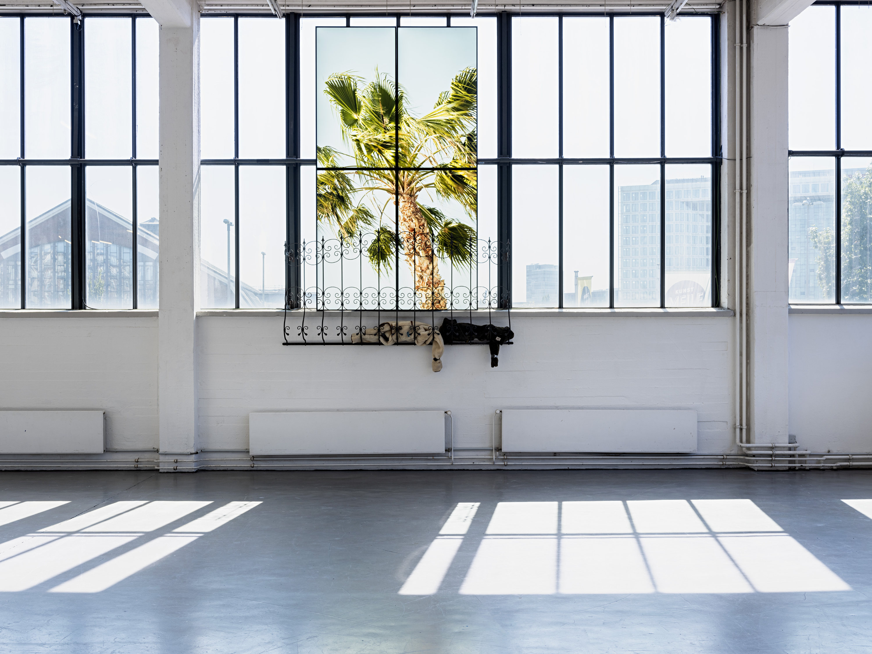 An image of an art installation in an industrial-style gallery space with large floor-to-ceiling windows with black metal frames. The central windowpane contains 4 video screens showing the image of a palm tree with green fronds against a bright sky. Below the video screens, a decorative wrought iron balcony railing is mounted on the wall.