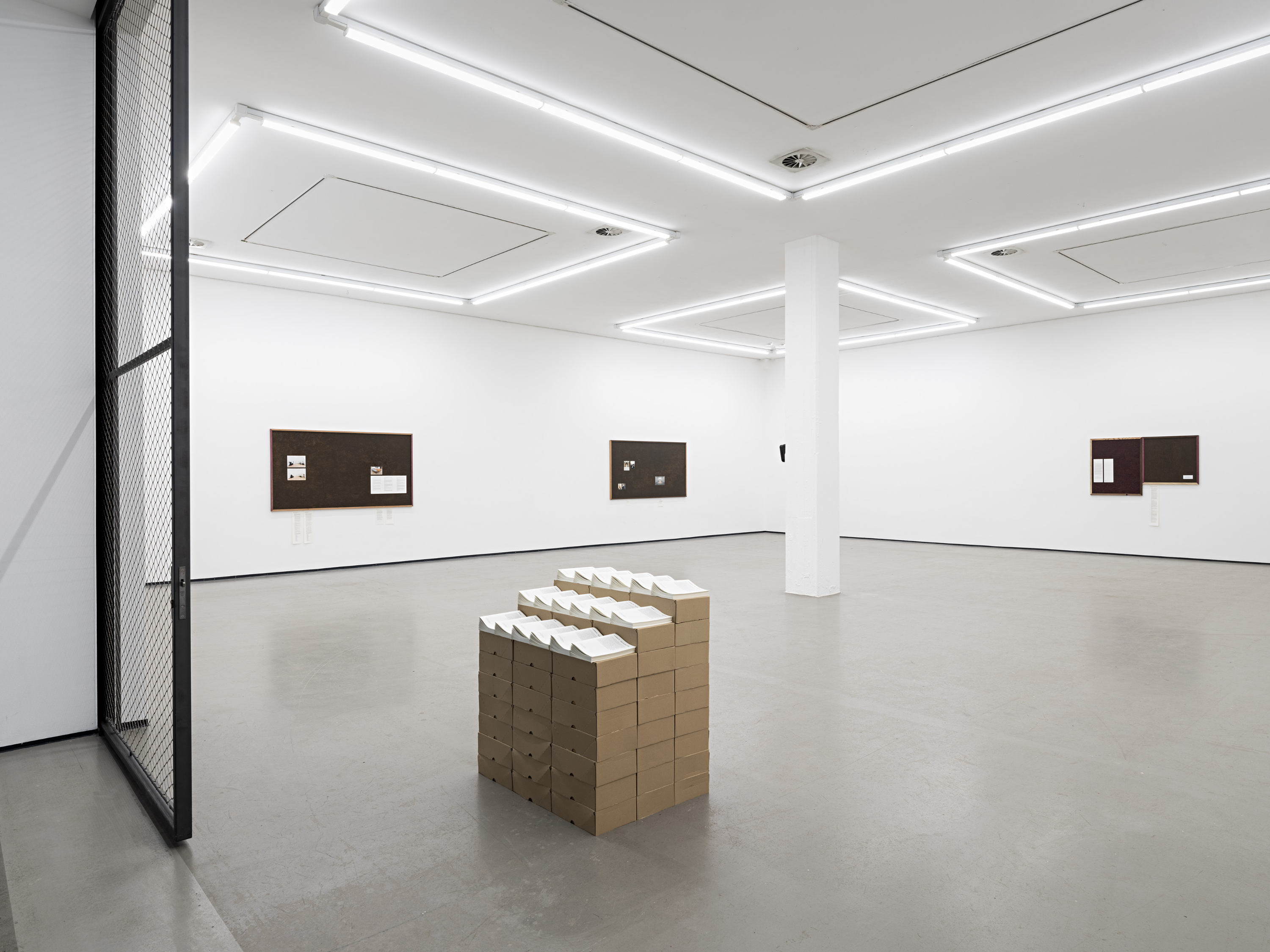 An image from the large, open doorway of a contemporary gallery with white walls and polished concrete floors . The ceiling feature four, large square light fixtures made from neon tubes. At the entrance to the room are three ascending stacks of brown-paper boxes, each stacker taller than the next. On top of each stack is an organized array of white paper pamphlets. On adjacent gallery walls, three art works of hung consisting of dark brown chipboard.
