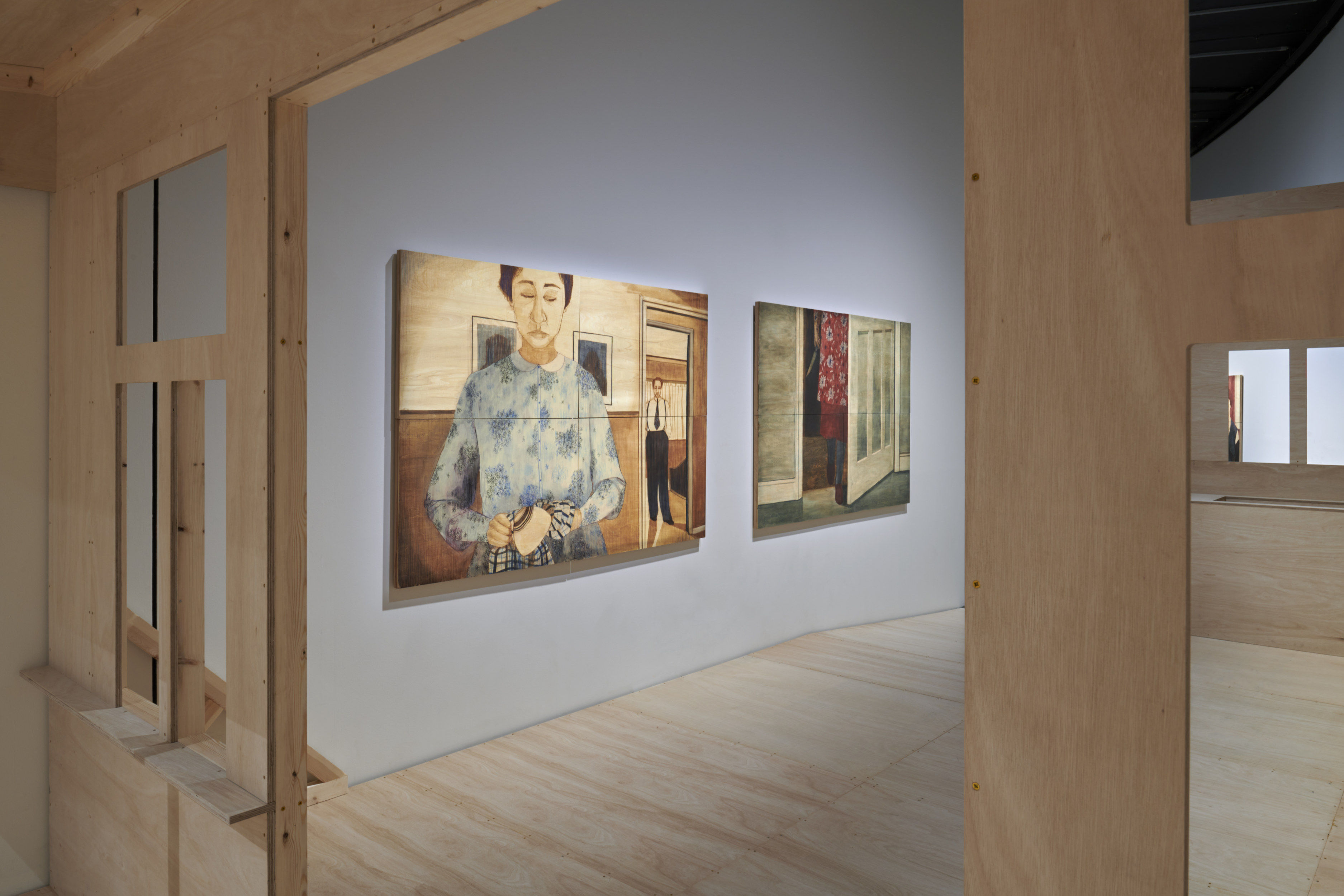 A contemporary art gallery shows two large paintings on an wall to the left. At the foreground of the image is a wall made of light wood, in whuch doors and windows have been cut so that the gallery is visible.