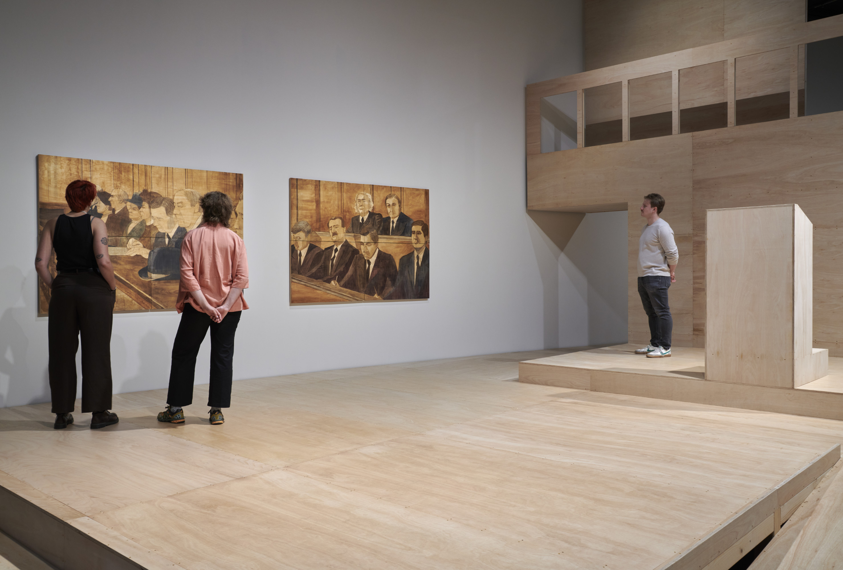 Three people observe paintings of historical figures in a modern art gallery with wooden flooring and minimalistic design.