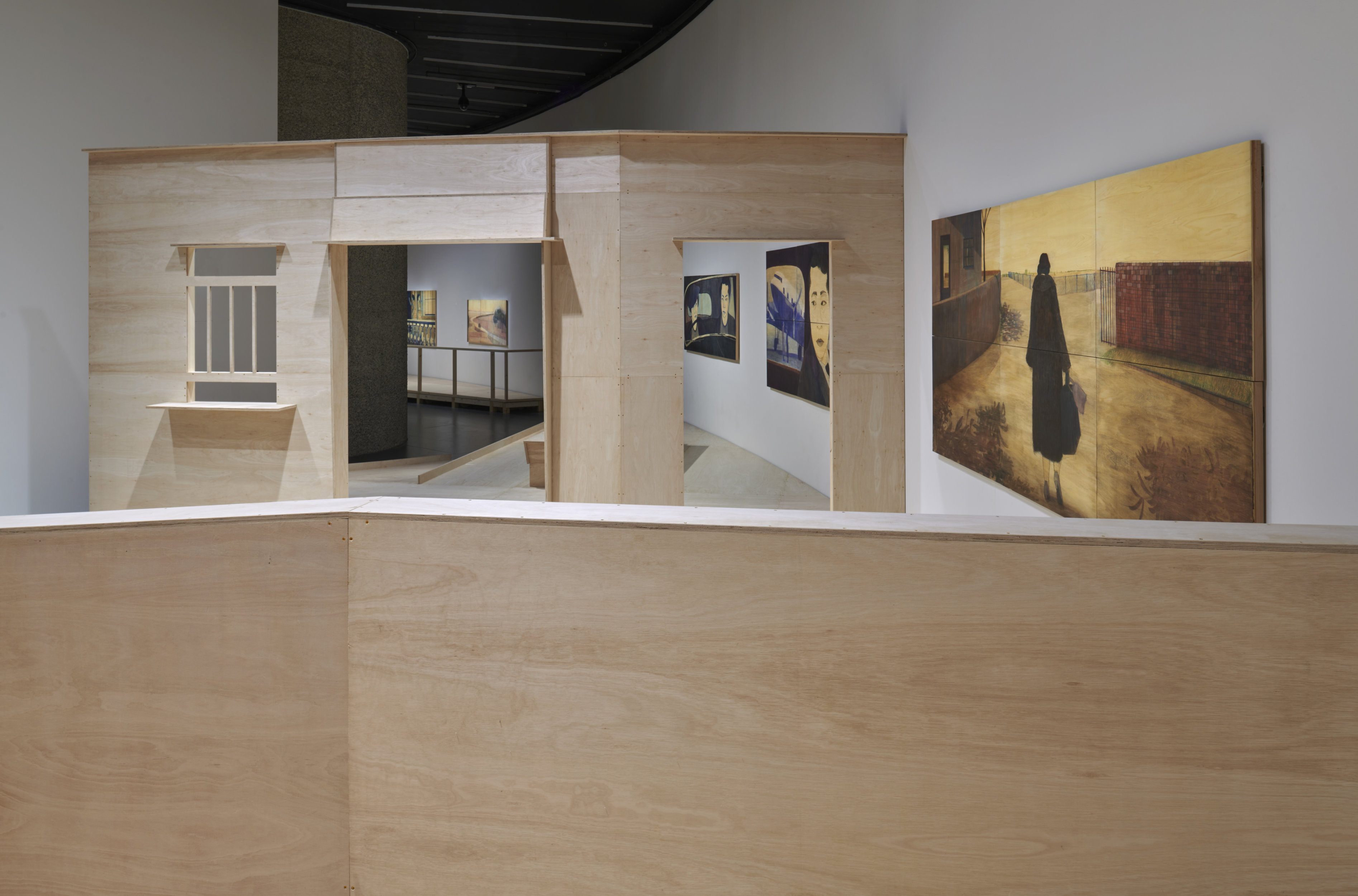 A contemporary art gallery features elements of a domestic stage set in minimalistic light wood. The facade of a house with window to the left and two door frames divide the space. To the right is a large painting with a yellow background, and a darkend figure at the center of the image depicted from behind. Through the wooded stage set, four large paintings can be seen. In the foreground of the image, a low wooden wall.
