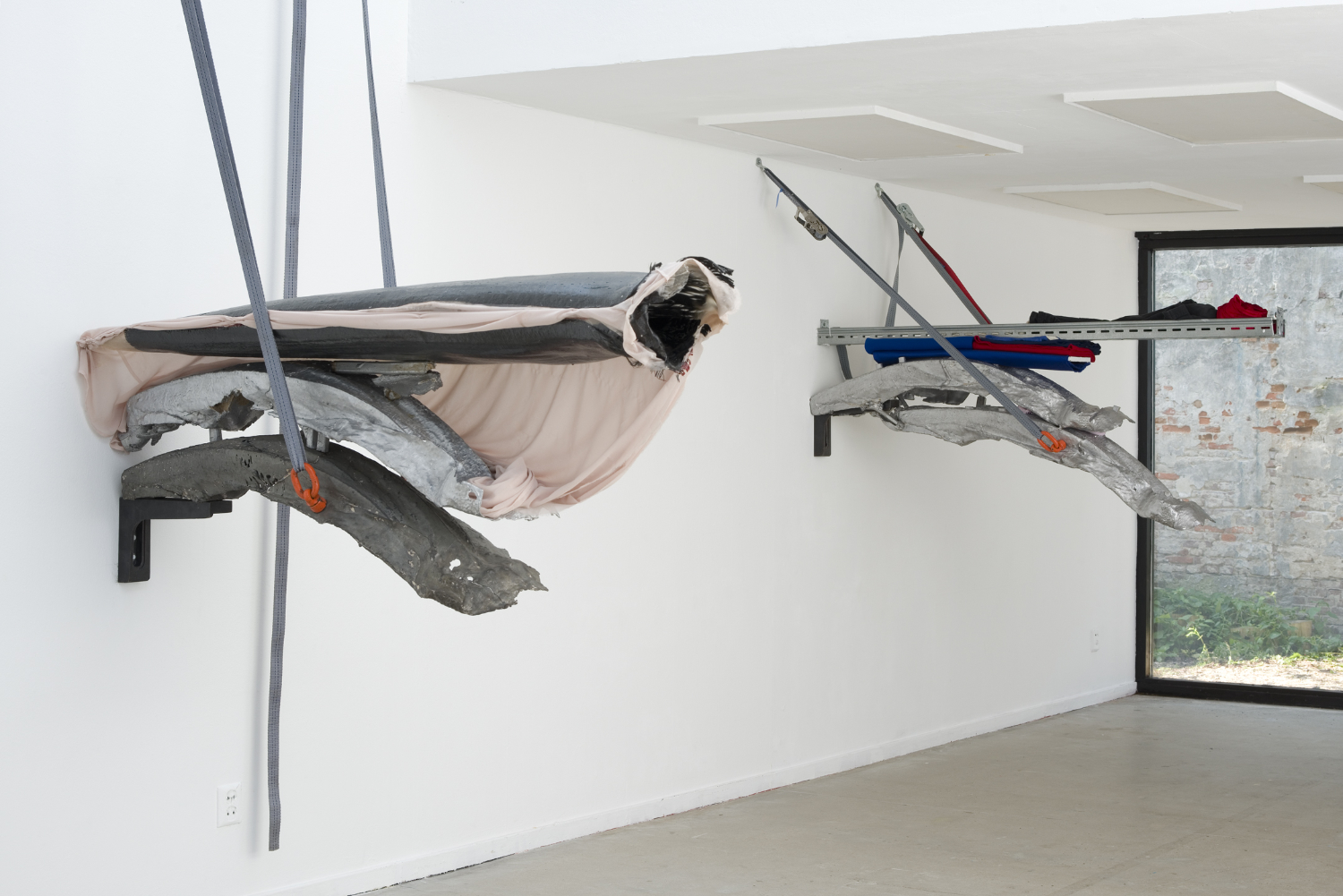 A contemporary gallery with two large sculpture suspended out of view from the ceiling and mounted against the wall. The sculptures are made of stacked metal and textile that resemble bolts of fabric.