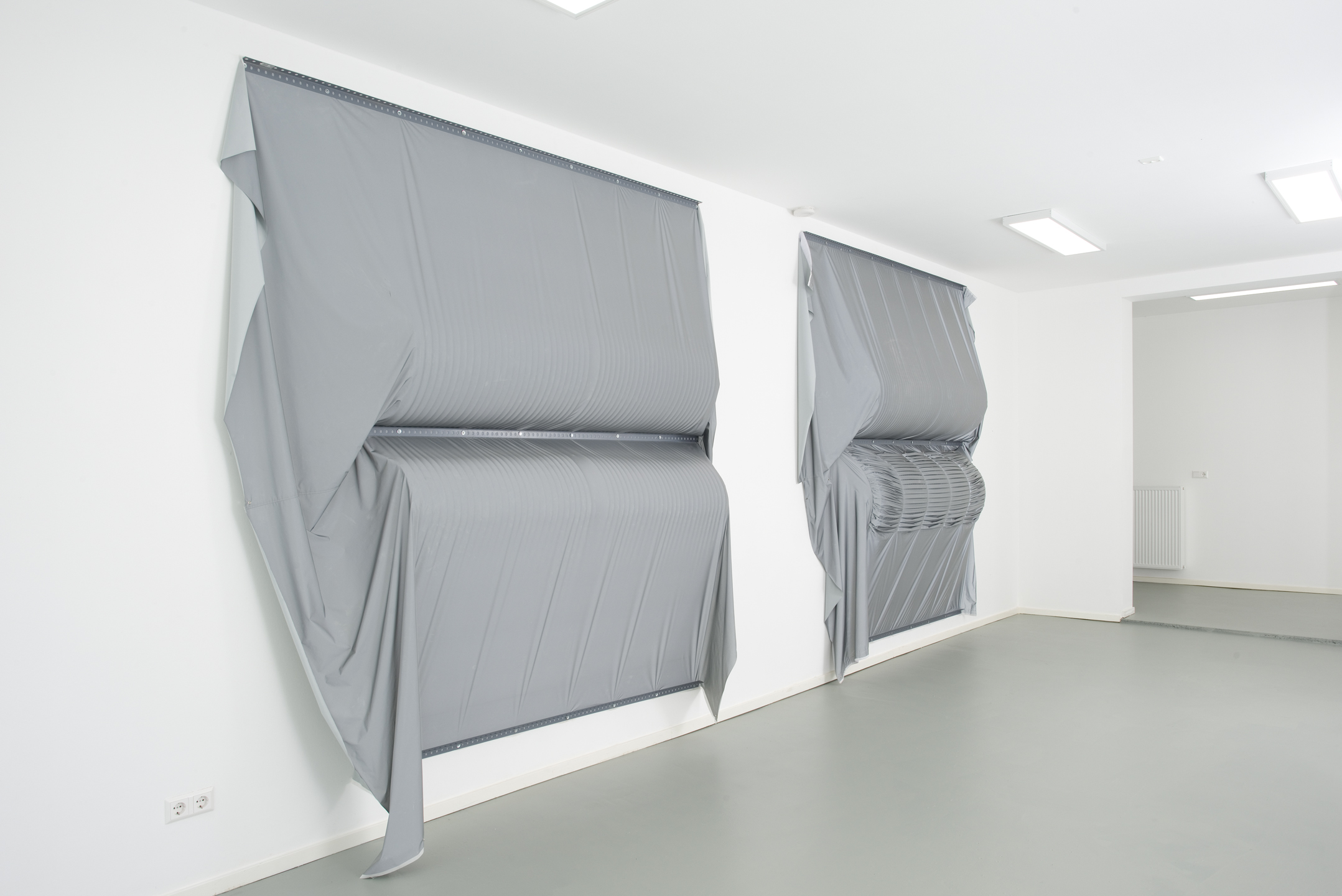 A contemporary art gallery with grey concrete floor and white walls. On the wall two large textile works, made of a grey iridescent, draped fabric are mounted.