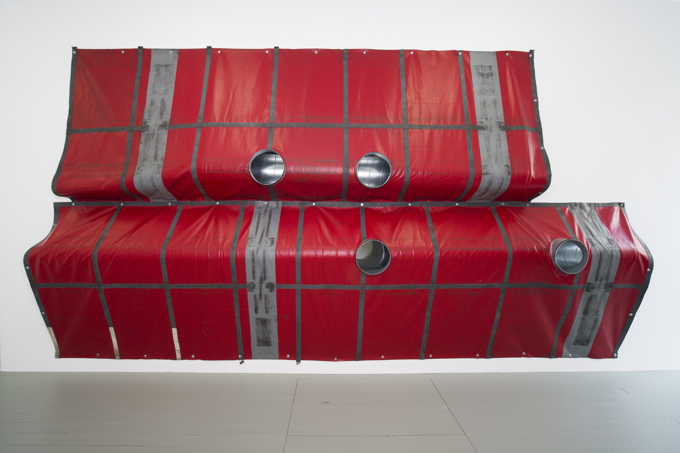 A red and gray industrial fabric installation with circular openings, displayed against a white wall.