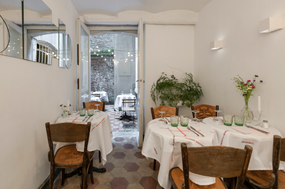 A small cafe with tile floor featuring three tables set for dinner and wooden chairs. Through a doorway, where fairy lights are strung, two more similar tables are visible.