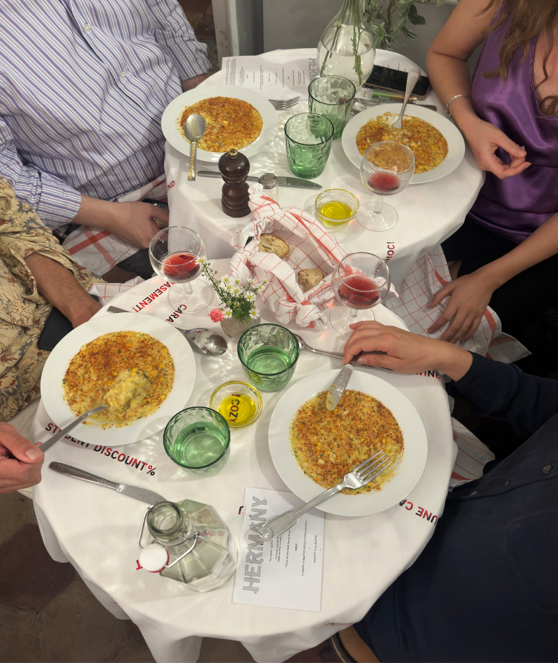 A close-up image, slightly from above, shows two circular tables, laid with a white tablecloth atop which sits a meal, glasses, and cups, Four people surround the table and are visible only until the shoulders.
