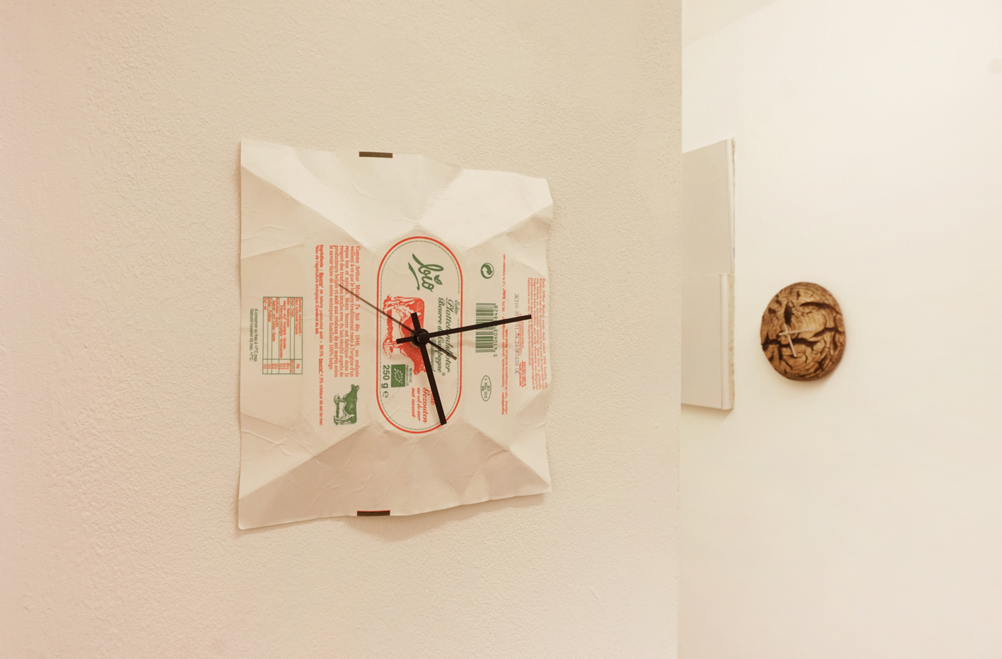 A white gallery wall, shows two works of art. In the foreground, s wall clock with hands attached to a wrinkled square piece of paper featuring various text and symbols. A circular carved clock, made from a loaf of bread is visible on an adjacent wall.