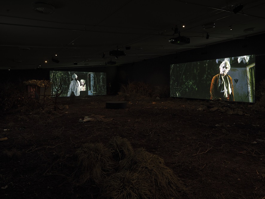 A pitch-black space shows two large screens with projections. On the left two figures enaged in conversation, wearing white wigs. On the right a figure faces forward, wearing a red jacket and a white wig.