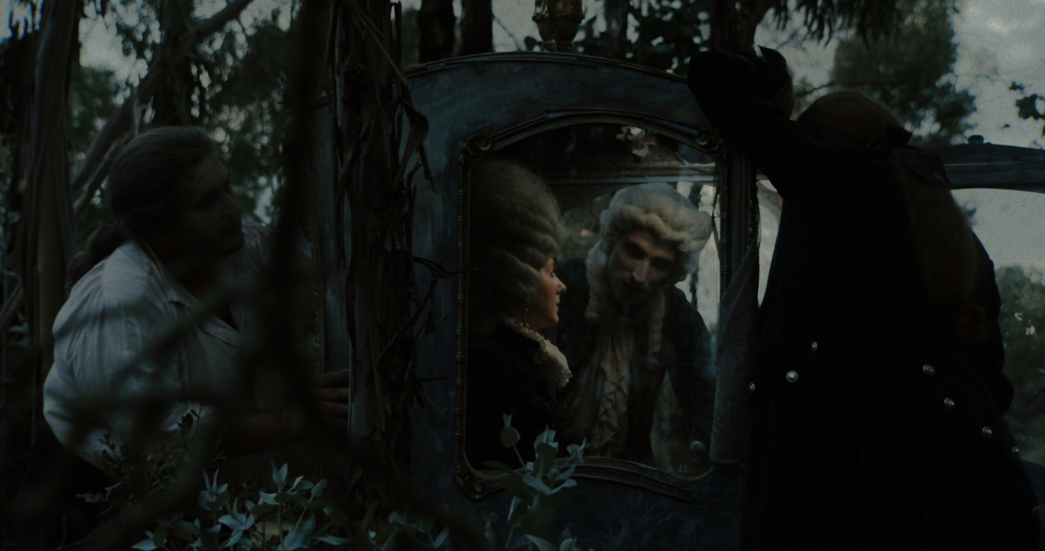 A darkened film still shows four figures in a nighttime forest wearing, positioned around an open carriage. The figures are dressed in costume from the 18th century with ruffled shirts and wigs. Two figures sit in the carriage and are engaged in conversation.
