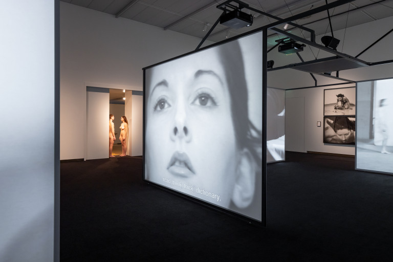 A contemporary gallery shows multiple screen hung from the ceiling featuring black and white images. The screen in the foreground shows the close-up face of a woman. In either side of the doorway to the left of the screens, two nude figures are visible leading into another gallery.