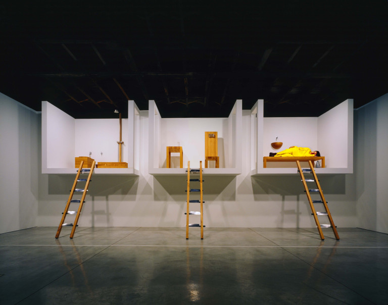 A contemporary gallery space with a dark ceiling and concrete floor. Built against a far wall are three separated and elevated platorms, enclosed except for the top. The left platform has a wooden shower and toilet, the middle platfrm has a table and high-backed wood chair, the right platform has a wooden bed atop which a figure in bright yellow is lying down. A ladder with wrungs made out of upturned knifes leans against each platform.