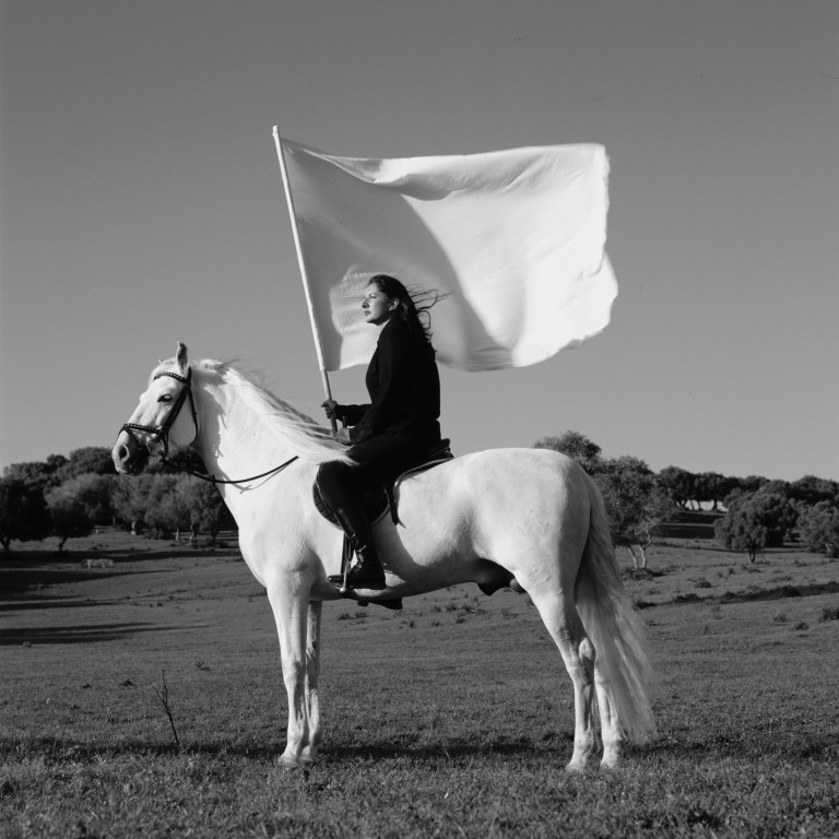 A black and white photoraph shows a rural landscape. In the foreground, a woman in black sits atop a white horse carrying a large white flag. The horse and woman are in profile and look into the distance.