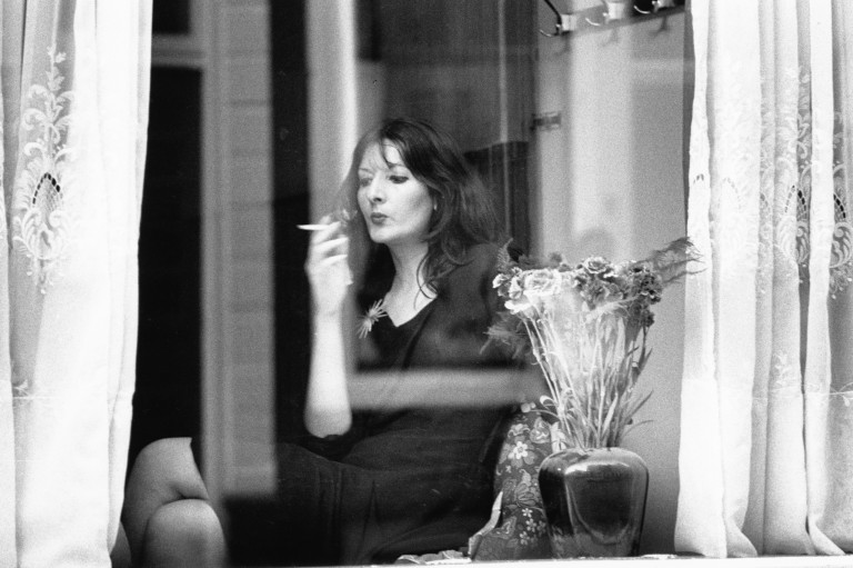 An archival photograph in black and white showing a seated woman behind a window in 3/4 profile smoking a cigarette. The woman is framed by lace curtains and a vase holding flowers sits in the window.