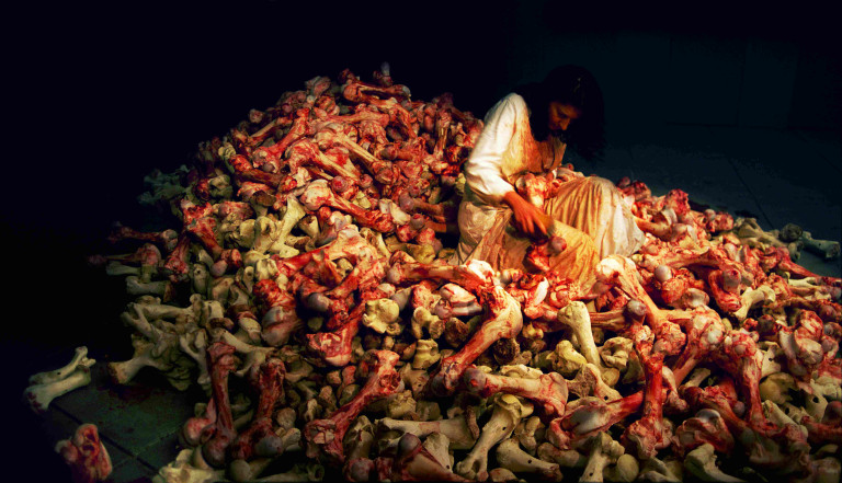 An image shows a darkened room with a large pile of bones, covered in blood. At the center of a pile a woman sits with a brush and a bone in her lap. She is wearing a long white dress also covered in blood and grime.