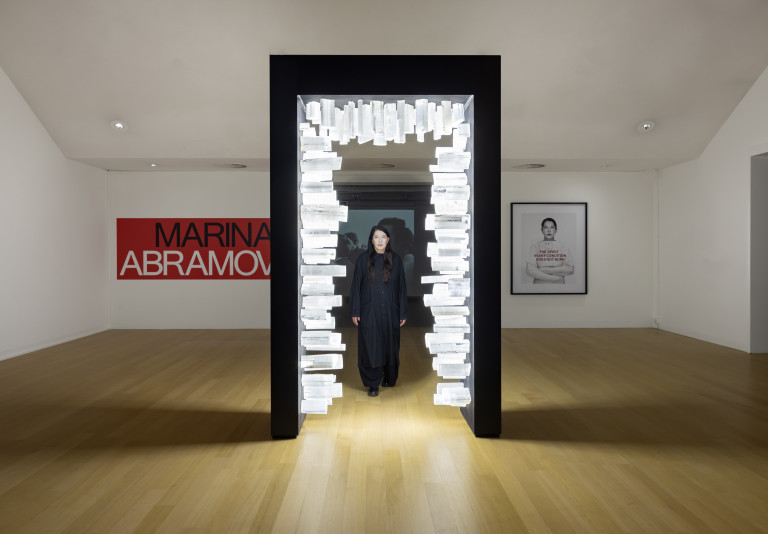 A contemporary gallery with light brown floor. In the center of the image a black doorframe, fitted on the inside with illuminated, plasticine shapes. A woman is framed by the door. On the far wall on the left of the image a red poster with black and white letters spell "MARINA ABRAMOVIC" on the right, a large photographic image framed in black shows a woman in white with red letters across the image.