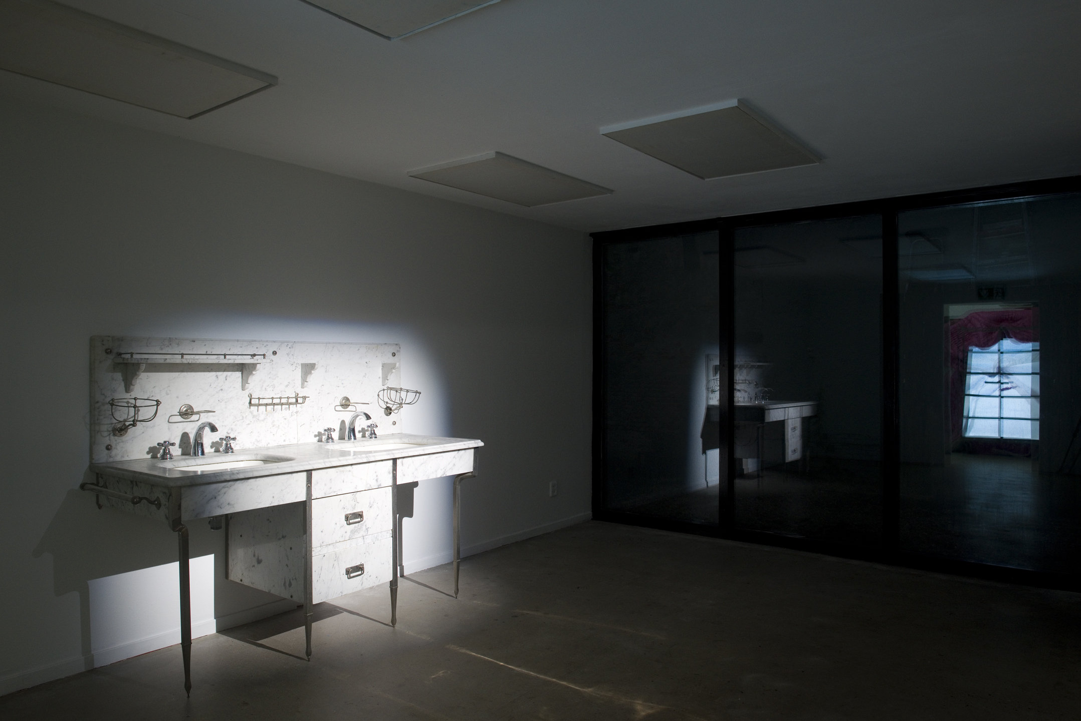 A contemporary gallery is dark except for a spotlight on a old-fashioned marble sink secured to a wall on the left. The sink includes two basins and metal wire sixtures for soap. The reflection of this sink can be seen in a darkened window on the right.
