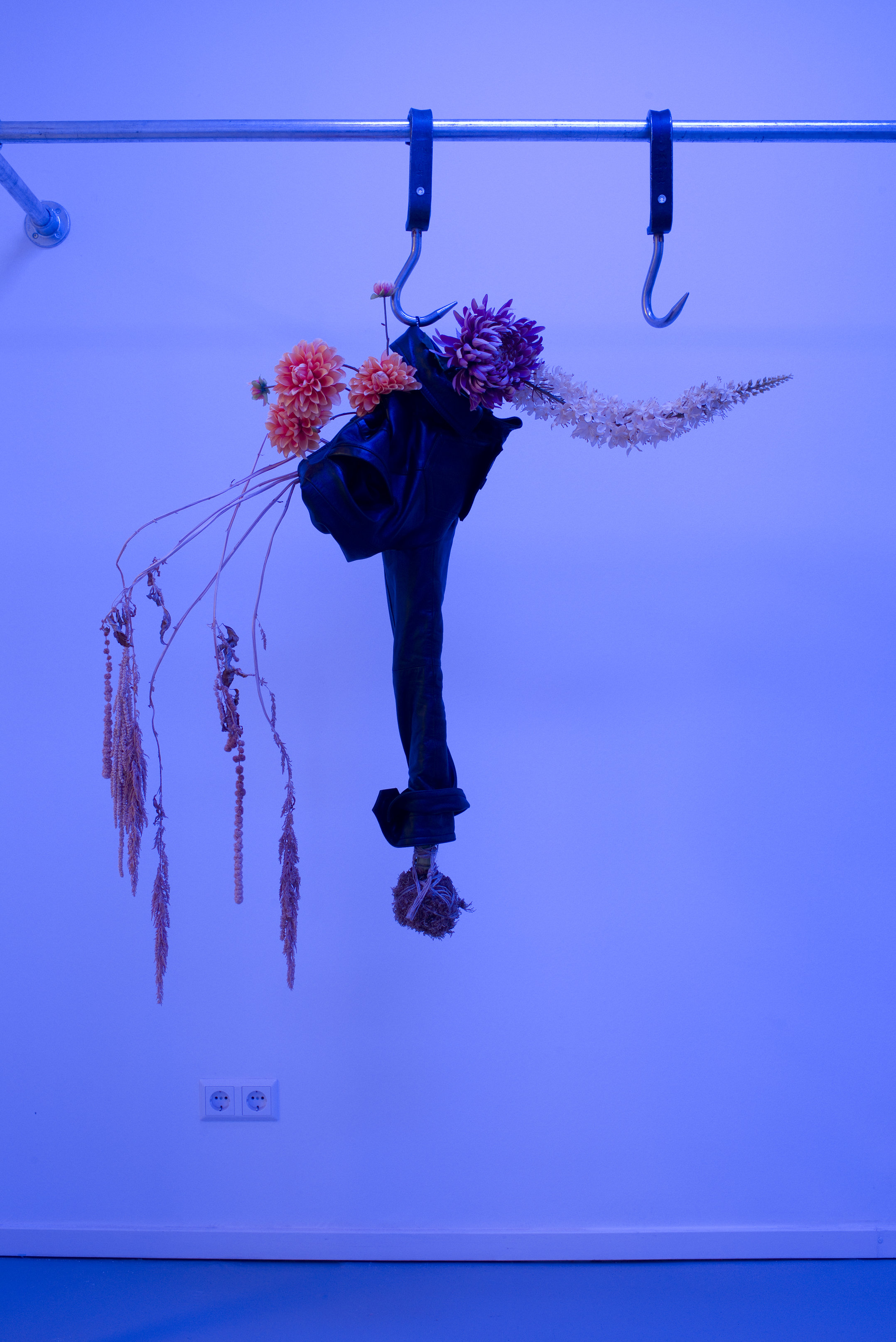 A close-up on a an art work in a contemporary gallery. A bouquet of dried flowers hangs from a long hook from a metal bar. Longer tendrils from the bouquet hang out of a paper wrapper. The room is cast in a blue light.