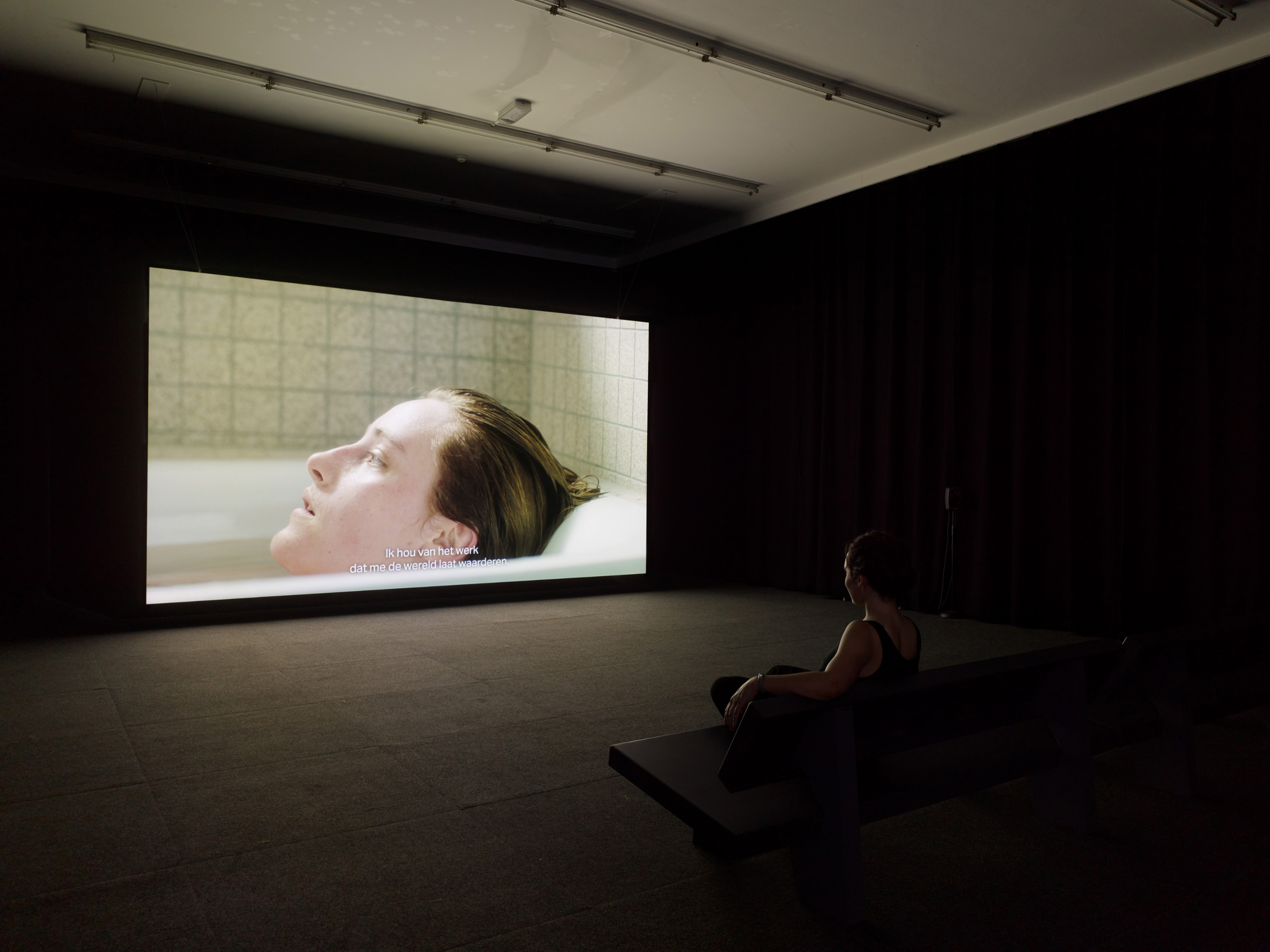 A darkened contemporary gallery with a large video screen. The screen shows a figure reclining in a bathtub with only their head visible.