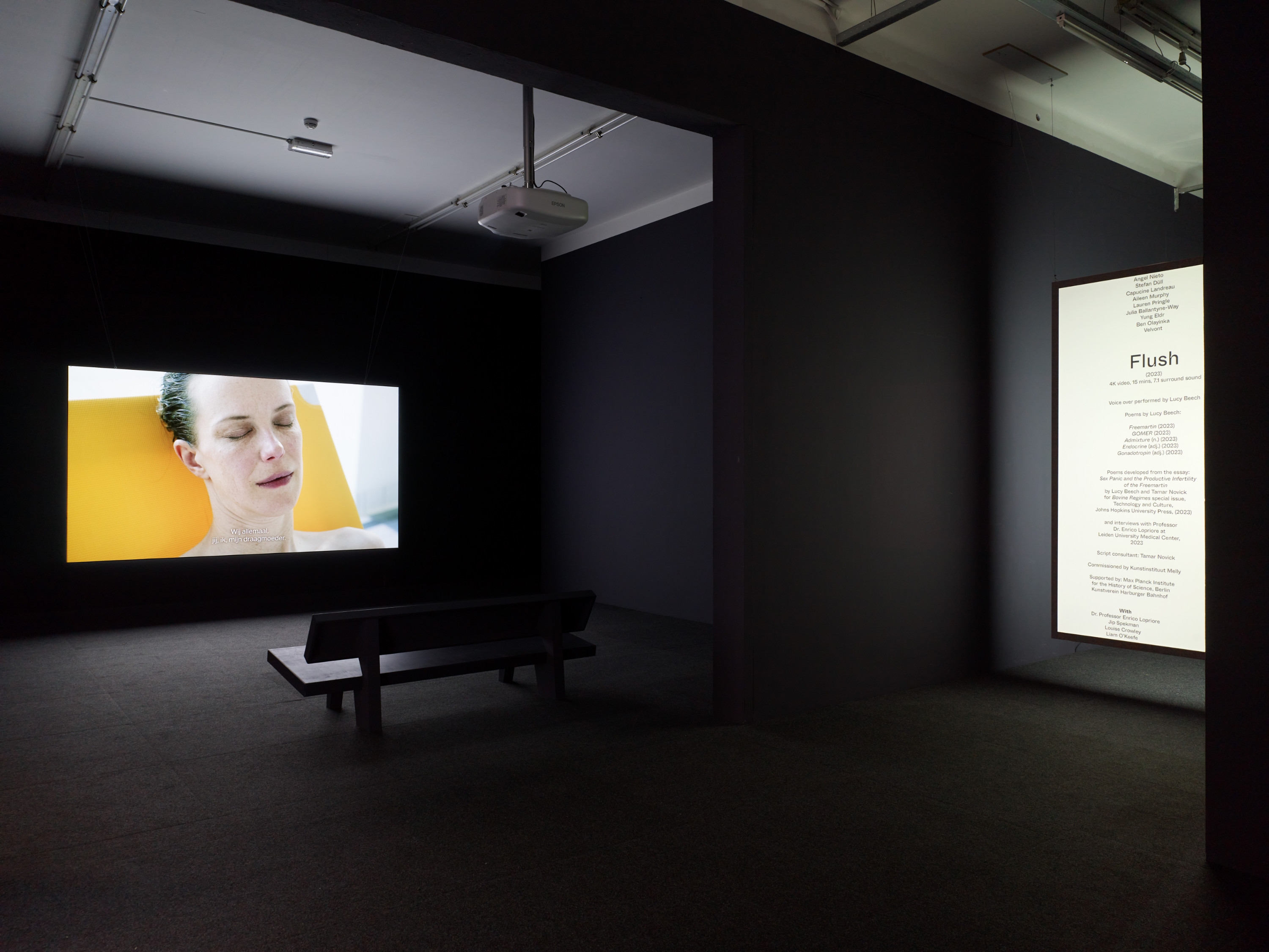 A darkened contemporary gallery with a large video screen. The screen shows a figure captured from the shoulders up with eyes closed. On the far right wall a screen with text. A bench is in the center of the space oriented toward the first screen.