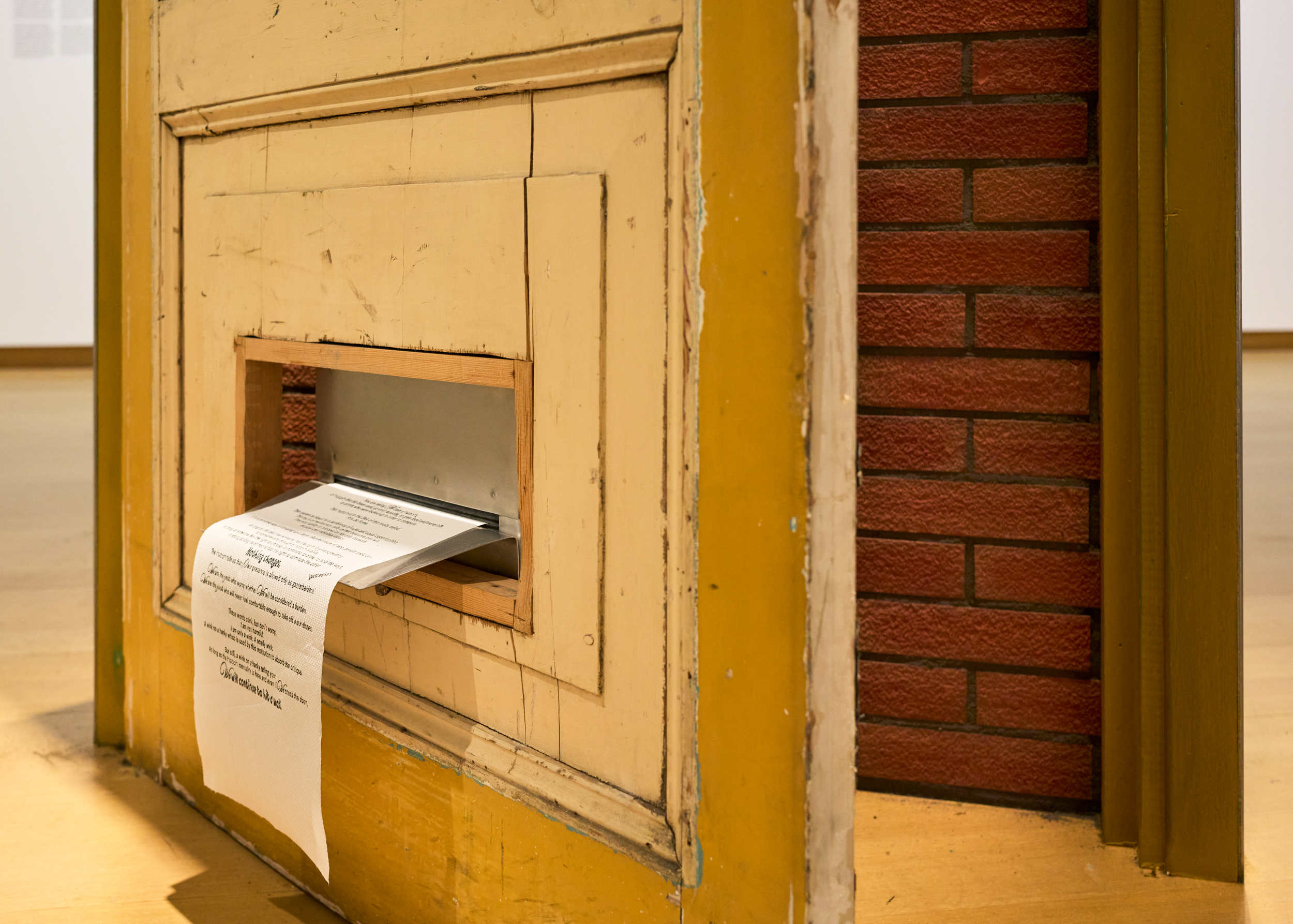 A close-up image of an art installation features an old wooden door with peeling yellow paint and visible wear. The door has a rectangular mail slot with a metal flap, through which a printed sheet of paper is protruding. The text on the paper is partially visible. Behind the door, there is a red brick wall, contrasting with the worn wooden surface. The installation is displayed in a gallery setting with light wooden flooring and a white exhibition wall visible in the background.