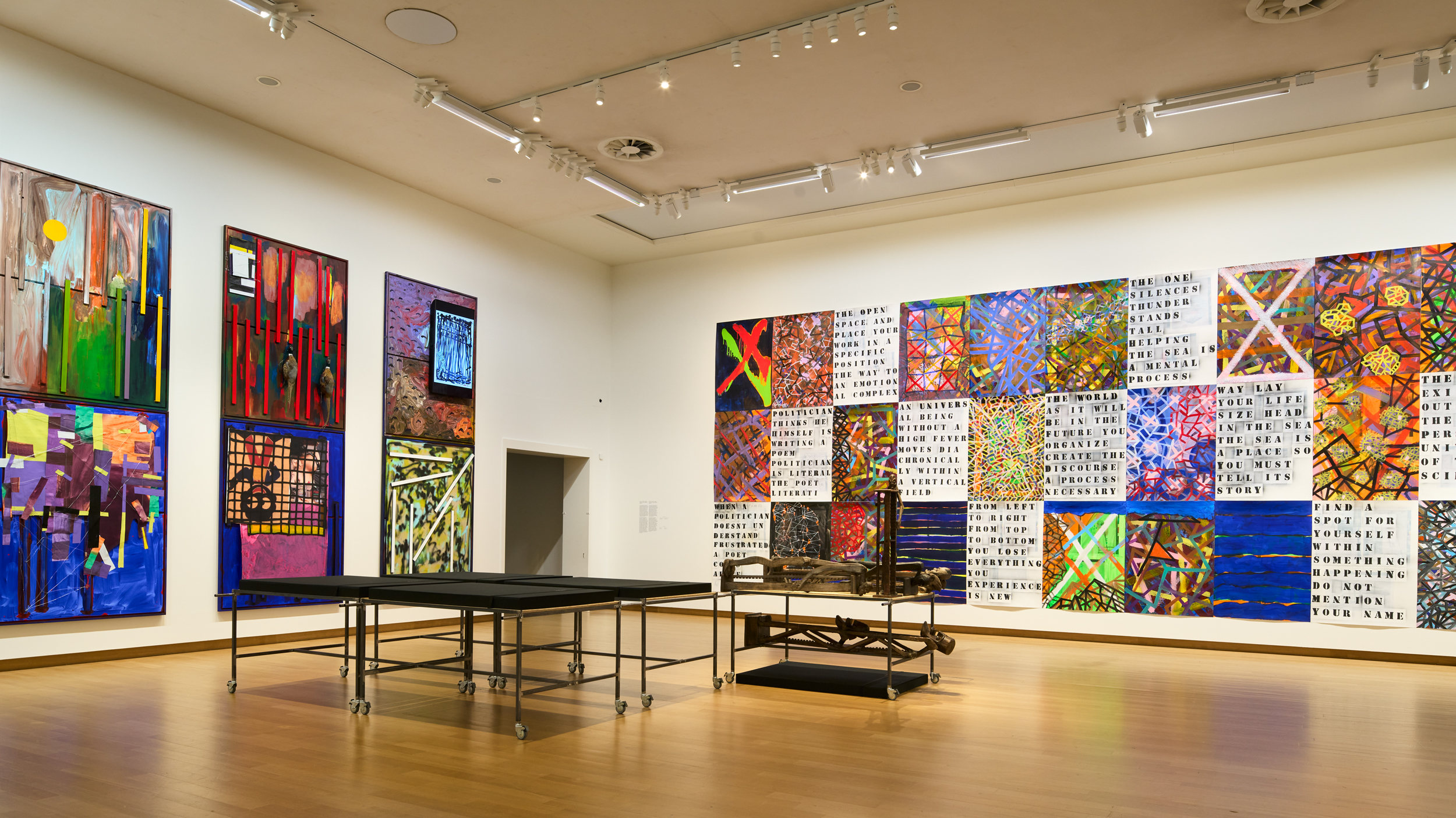 A large contemporary art gallery with wood floor. The walls are hung with an alternating grid of large, colorful, abstract paintings and white canvases with text . In the foreground